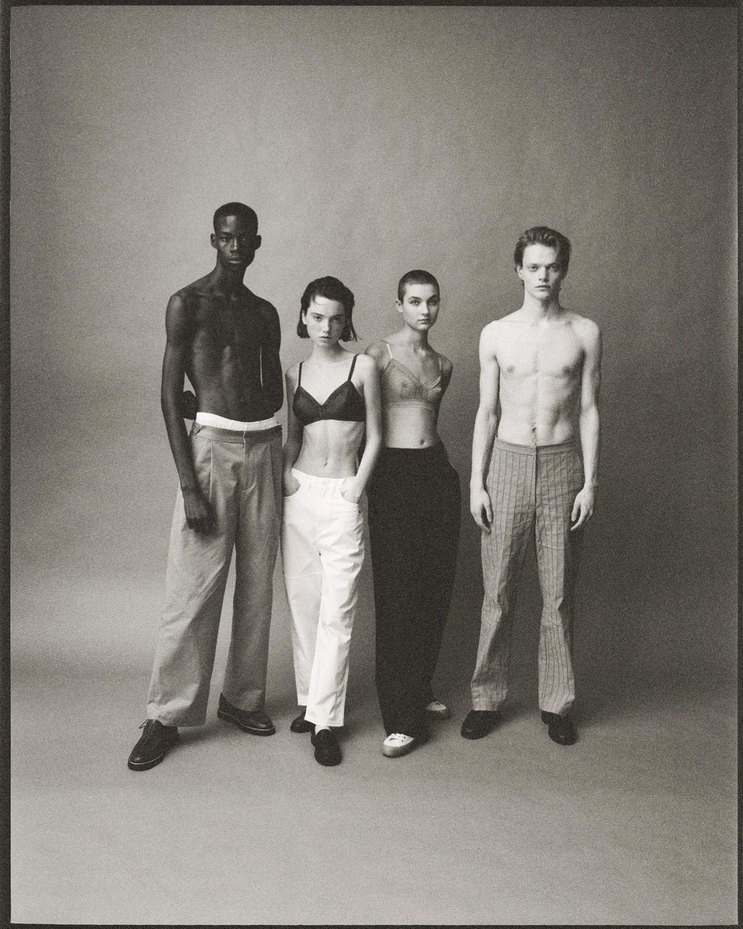 Four fashion models pose indoors against a grey studio backdrop. Two men are bare-chested, alongside two women in bras and trousers.