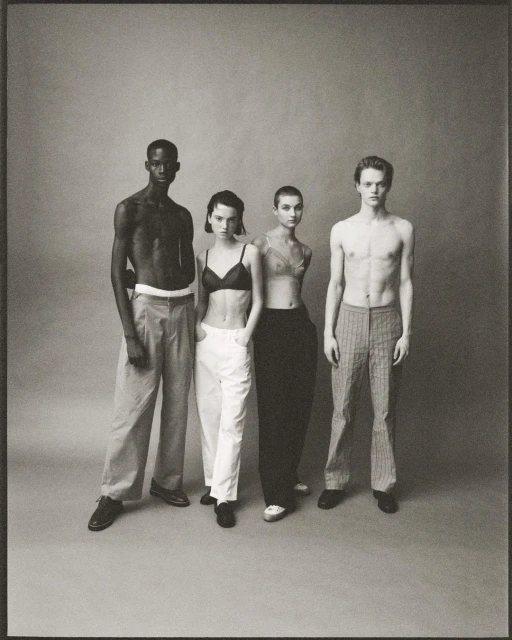 Four fashion models pose indoors against a grey studio backdrop. Two men are bare-chested, alongside two women in bras and trousers.