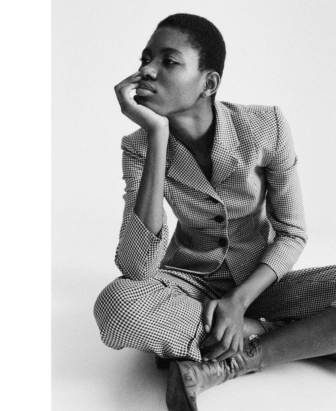 Black and white portrait of a Black woman in a checkered suit sitting cross-legged, with her hand on her chin, gazing to the left.