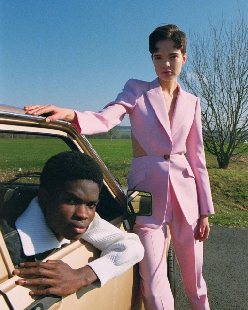 Two fashion models pose in a field beside a vintage car. One wears a pink pantsuit, the other wears a white sweater and black jacket.
