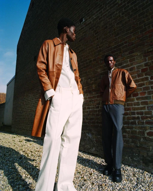 Two adult men pose in stylish outerwear against a brick wall, one in a long leather coat and the other in a brown leather bomber jacket.