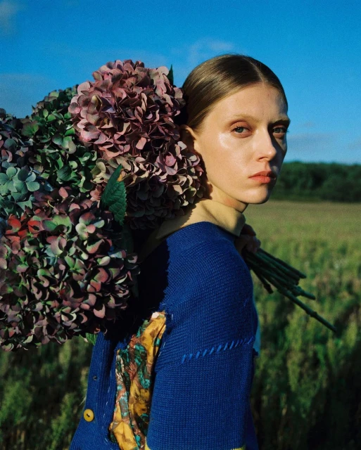 Woman poses with a large purple and green hydrangea bouquet in an outdoor field beneath a blue sky, wearing a blue knit sweater.