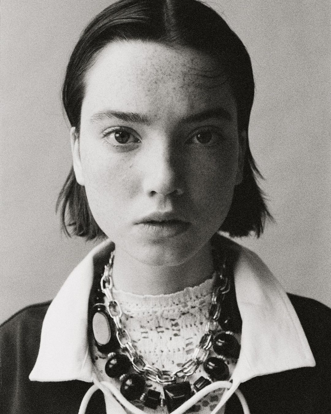 Headshot of a young woman with freckles and short dark hair, wearing a statement necklace, in a close-up studio portrait.