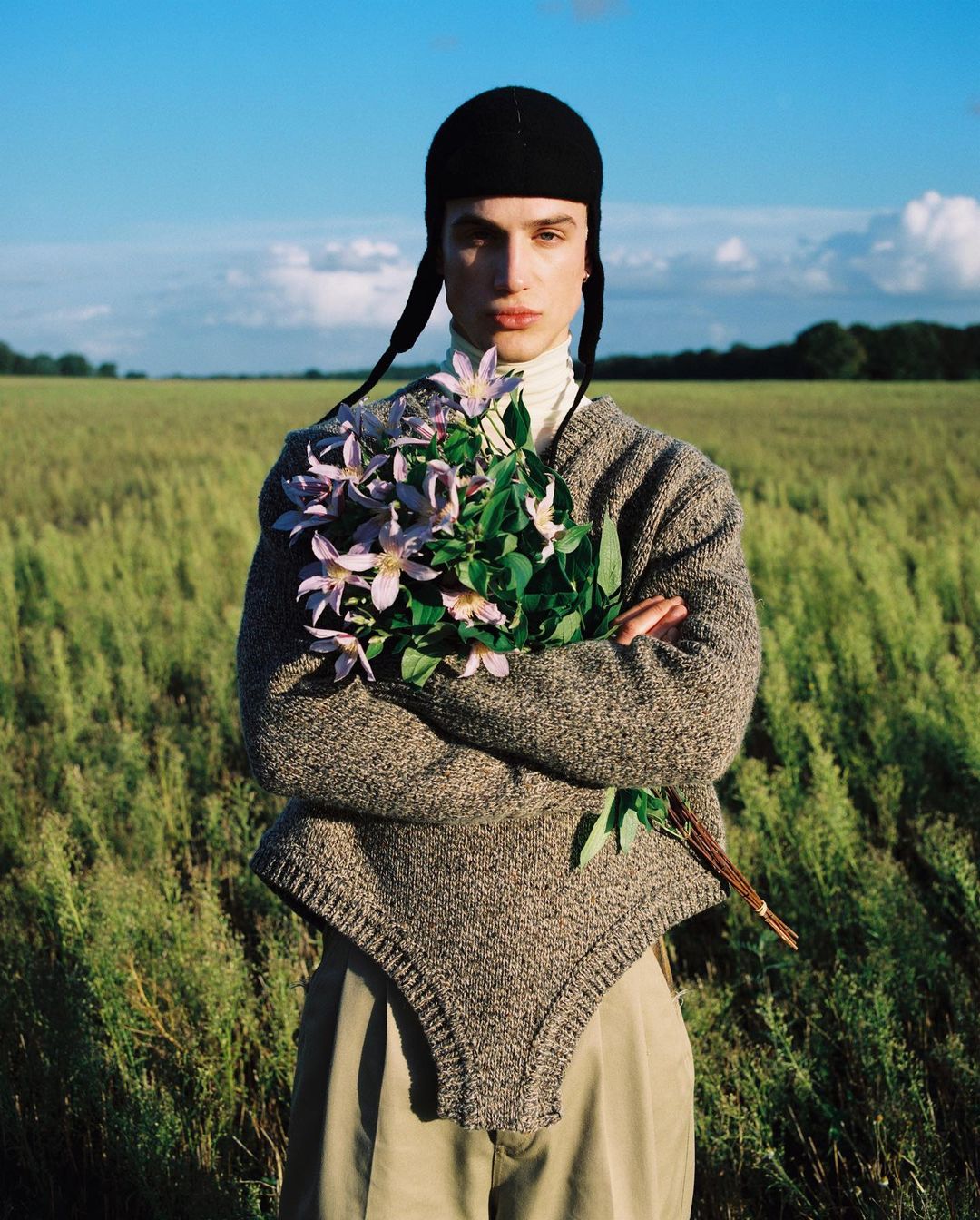 A person in a black hat and grey sweater holds a bouquet of purple flowers outdoors with a bright blue sky and green field background.