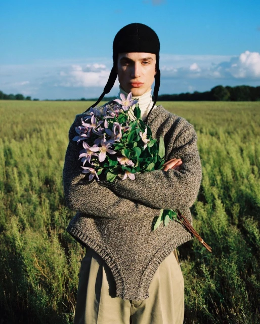 A person in a black hat and grey sweater holds a bouquet of purple flowers outdoors with a bright blue sky and green field background.