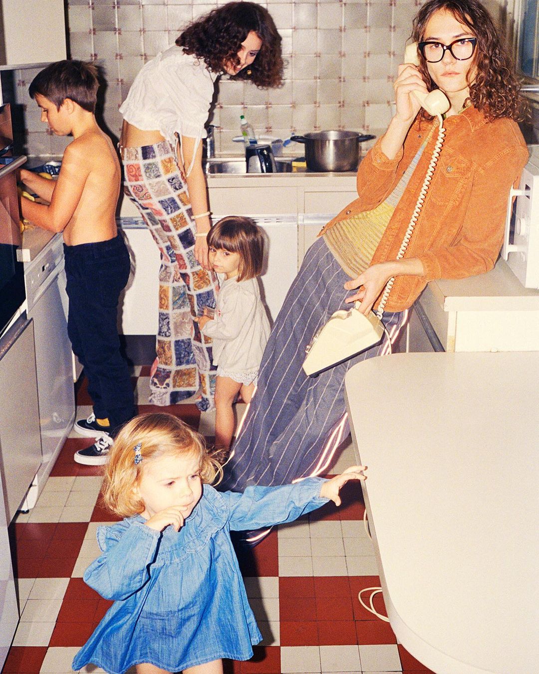 A group of children and adults are gathered in a brightly lit kitchen with a retro-style red and white checkered floor and various objects.