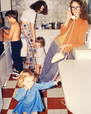 A group of children and adults are gathered in a brightly lit kitchen with a retro-style red and white checkered floor and various objects.