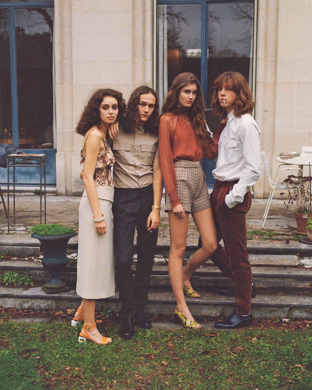 Four young adults pose on a stone patio in front of a grand stone house, showcasing vintage-inspired fashion in an outdoor setting.