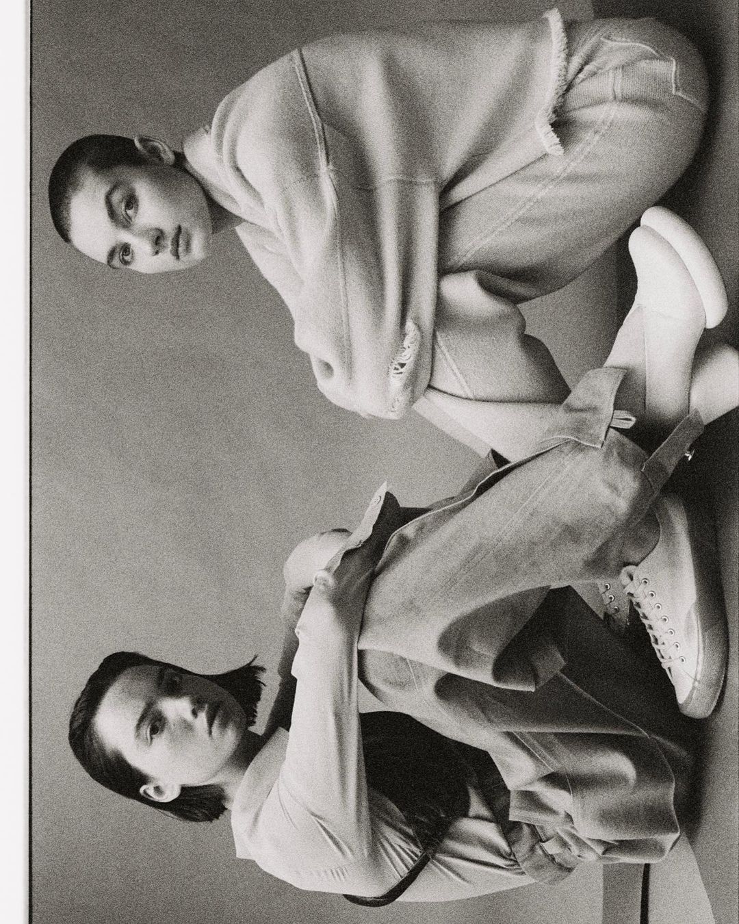 Two women in sweatshirts and jeans pose in a studio with muted grey background. The image utilizes a monochrome color palette.
