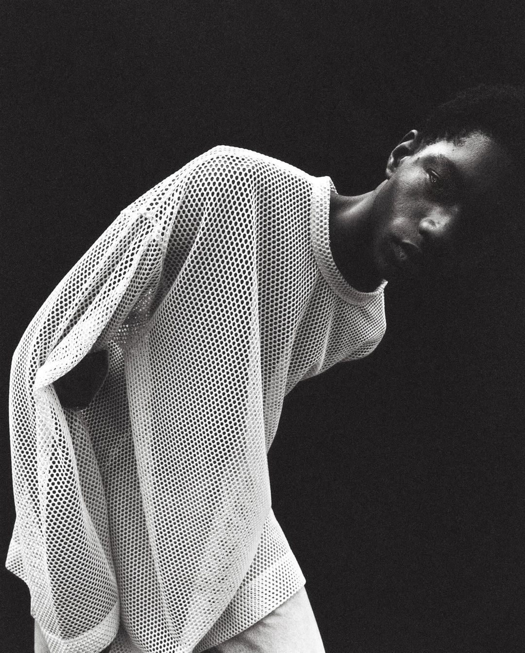 Black and white portrait of an adult Black man in a mesh shirt looking down with a dark background, medium studio shot.