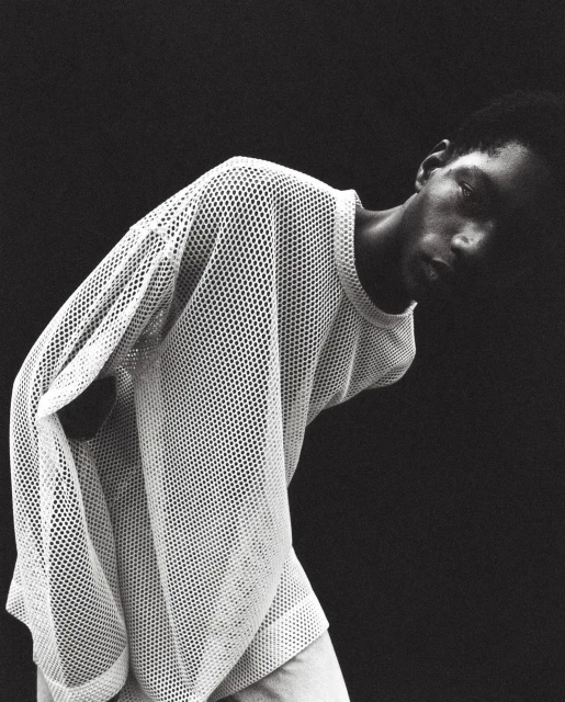 Black and white portrait of an adult Black man in a mesh shirt looking down with a dark background, medium studio shot.