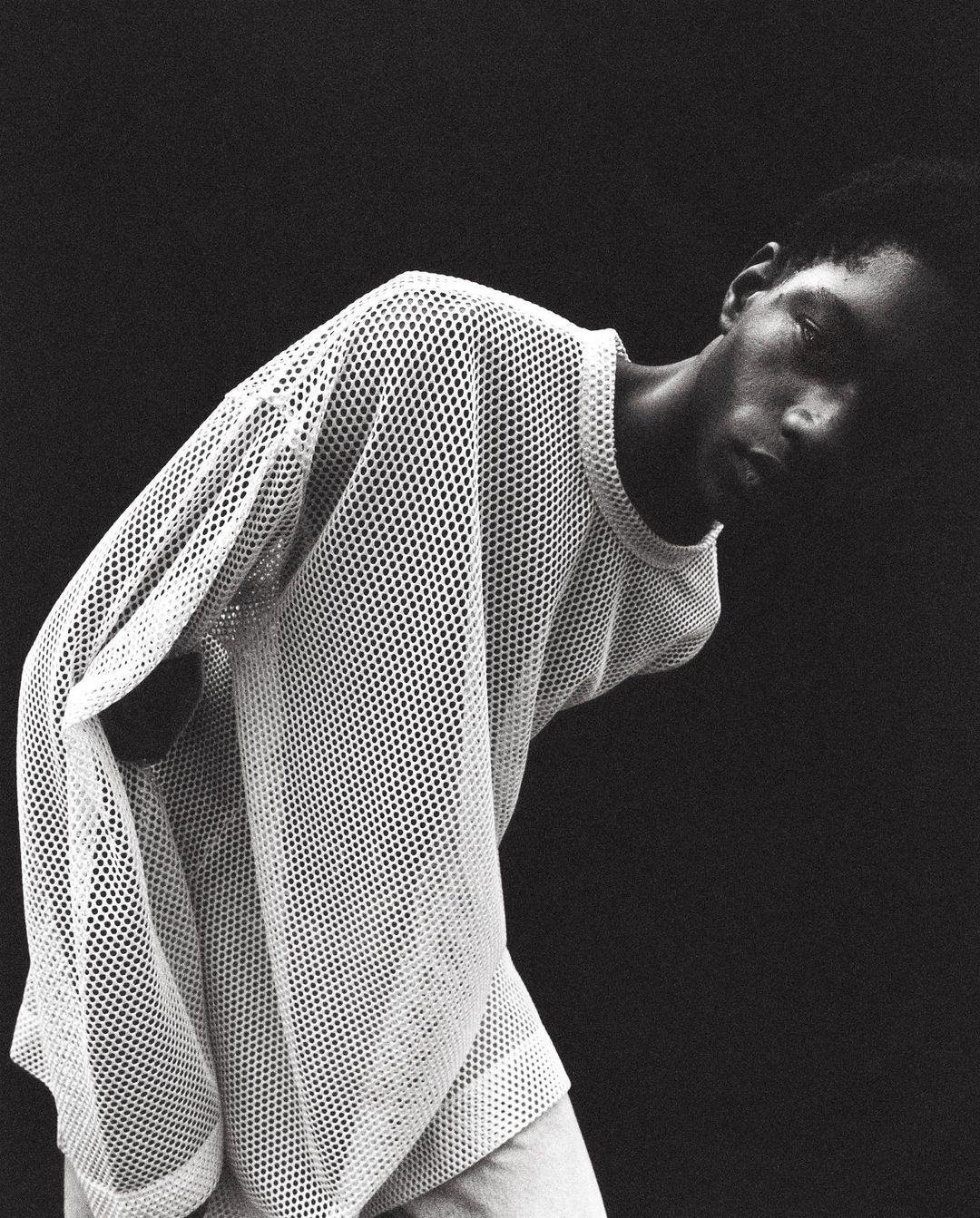 Portrait of a Black male model in a white mesh shirt against a dark background, showcasing fashion and studio photography.