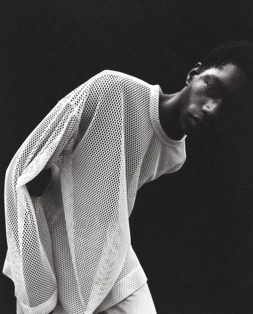 Portrait of a Black male model in a white mesh shirt against a dark background, showcasing fashion and studio photography.