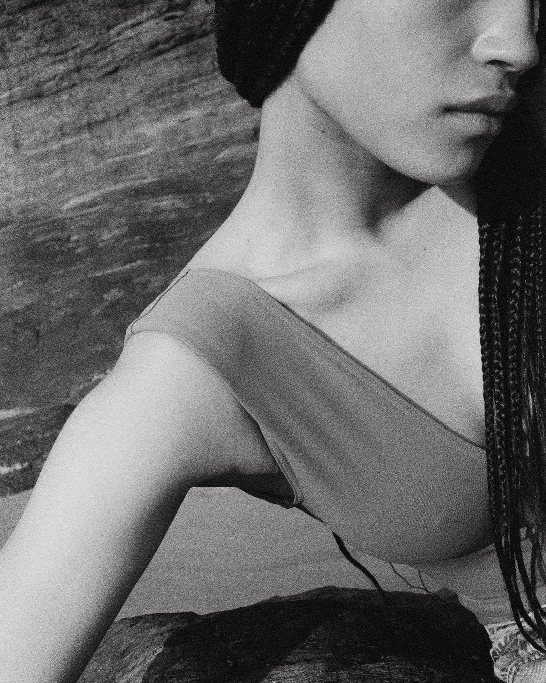 Monochrome close-up shot of an adult woman with braids and a knit hat posing near a rock formation with natural light and neutral tones.