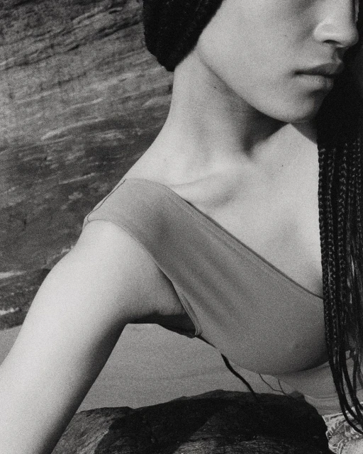 Monochrome close-up shot of an adult woman with braids and a knit hat posing near a rock formation with natural light and neutral tones.