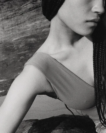Monochrome close-up shot of an adult woman with braids and a knit hat posing near a rock formation with natural light and neutral tones.