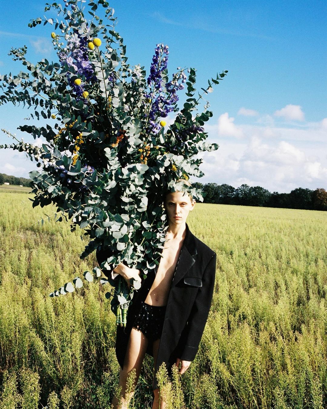 Male model posing with a large bouquet of flowers in a grassy field against a blue sky, wearing a black jacket and shorts, fashion.