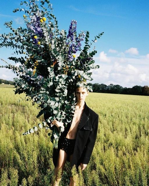 Male model posing with a large bouquet of flowers in a grassy field against a blue sky, wearing a black jacket and shorts, fashion.