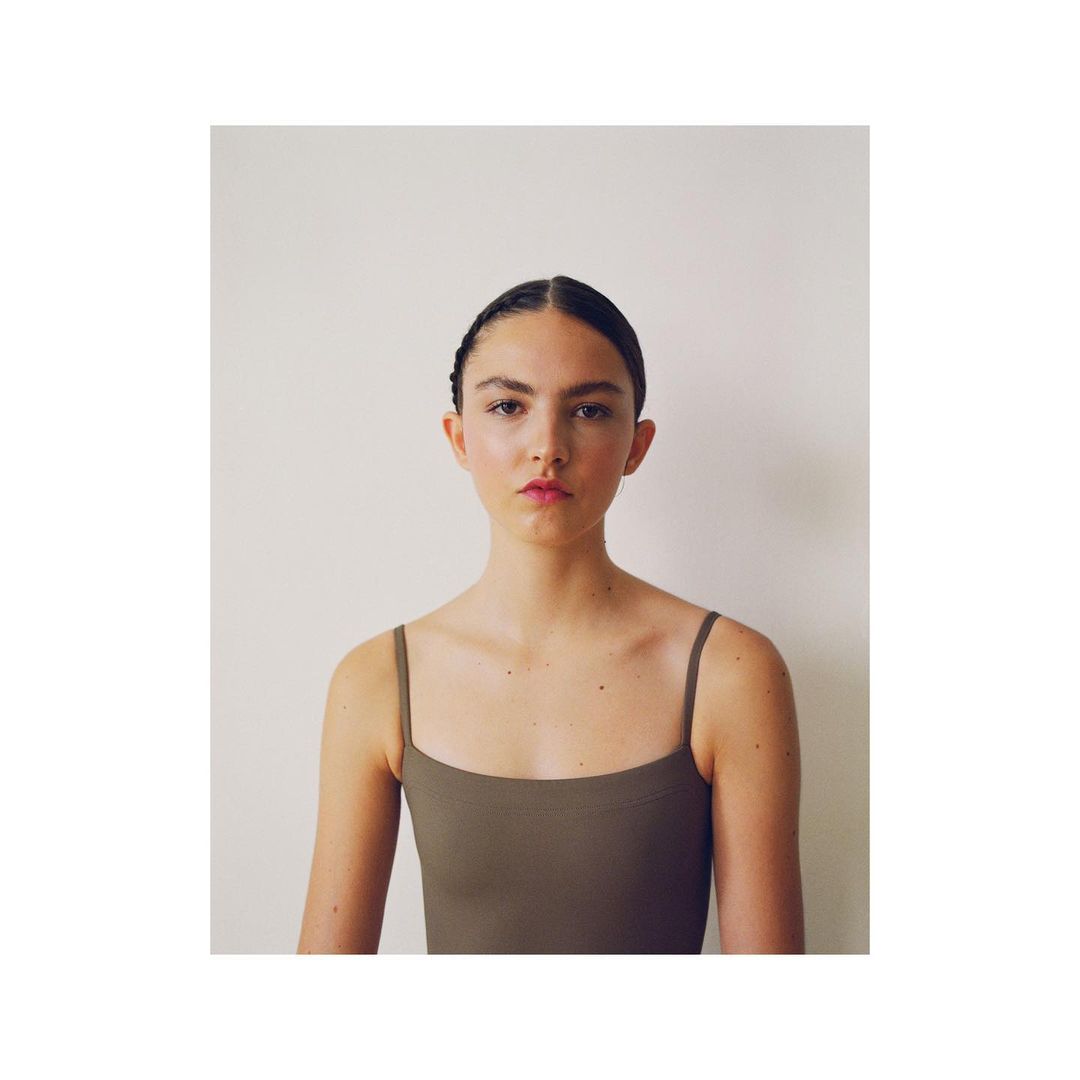 A portrait of a young woman with dark hair pulled back, wearing a simple brown top, against a plain white background in soft, natural light.