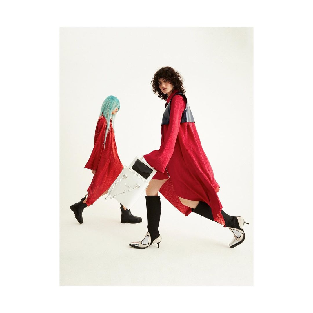 Two models in red dresses and boots pose in a studio against a white backdrop. One model carries a white box-like bag.
