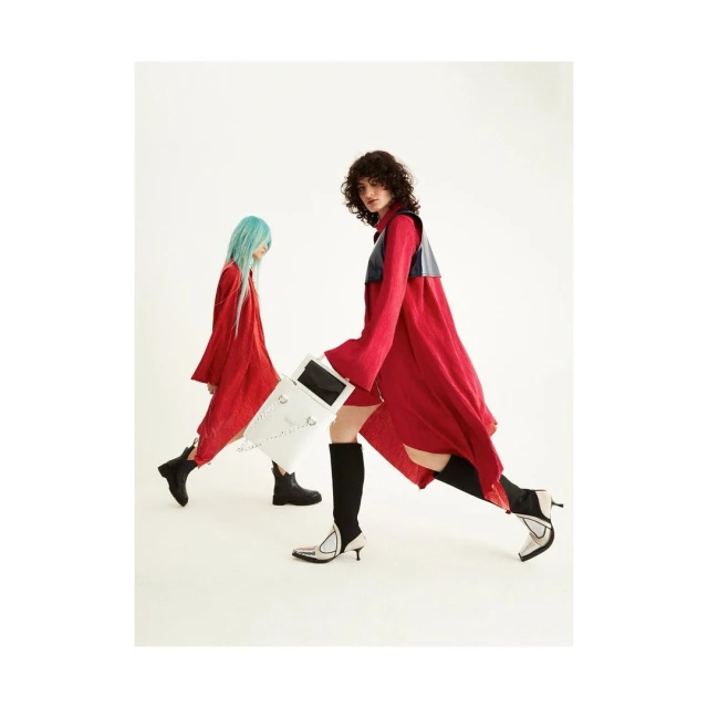 Two models in red dresses and boots pose in a studio against a white backdrop. One model carries a white box-like bag.