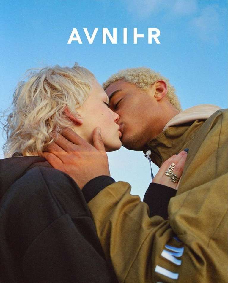 Close-up of a couple kissing in an outdoor setting under a clear sky. The couple has light blonde hair and are wearing fashionable clothing.