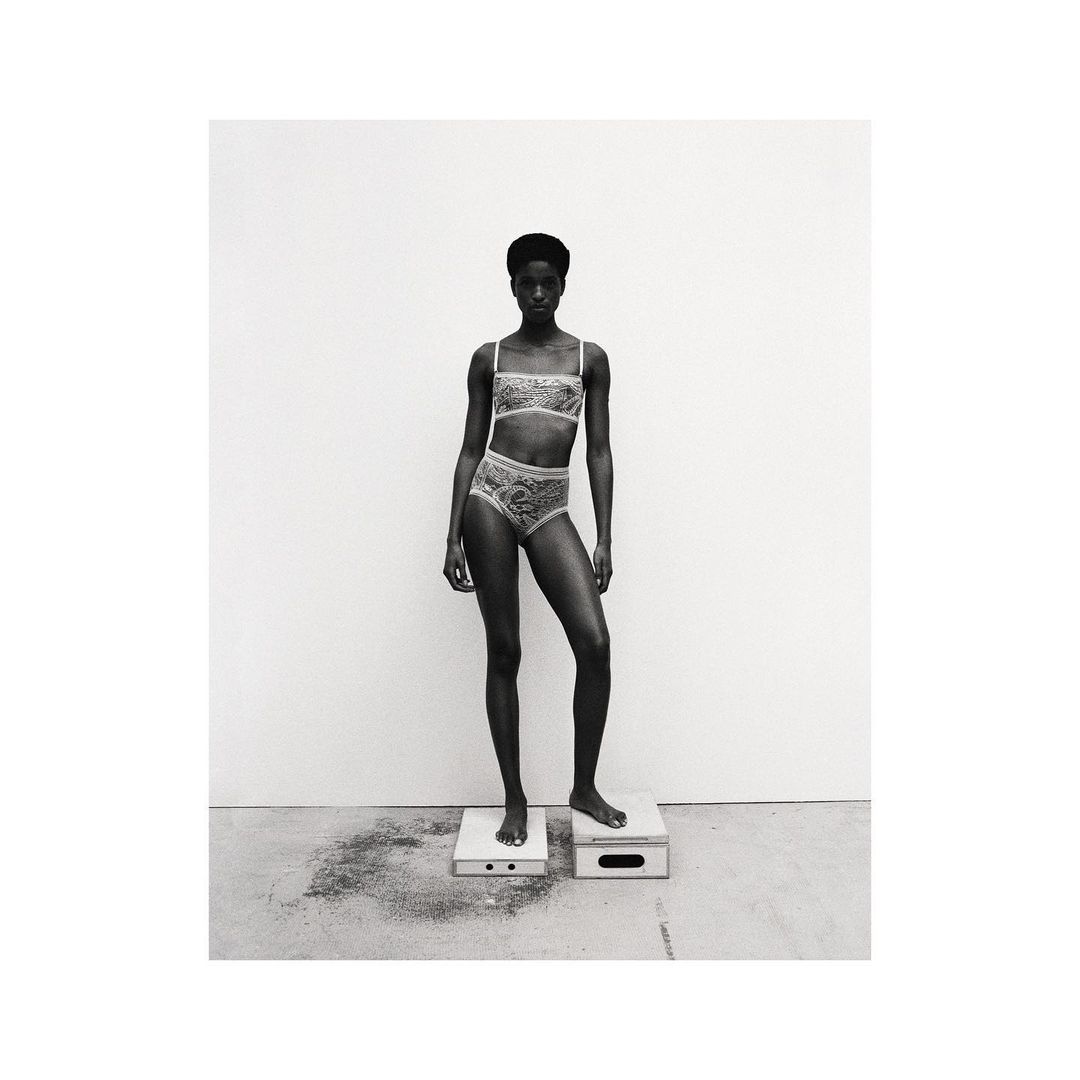 Monochrome studio shot of a slender Black woman in lace lingerie standing on wooden platforms against a neutral backdrop.