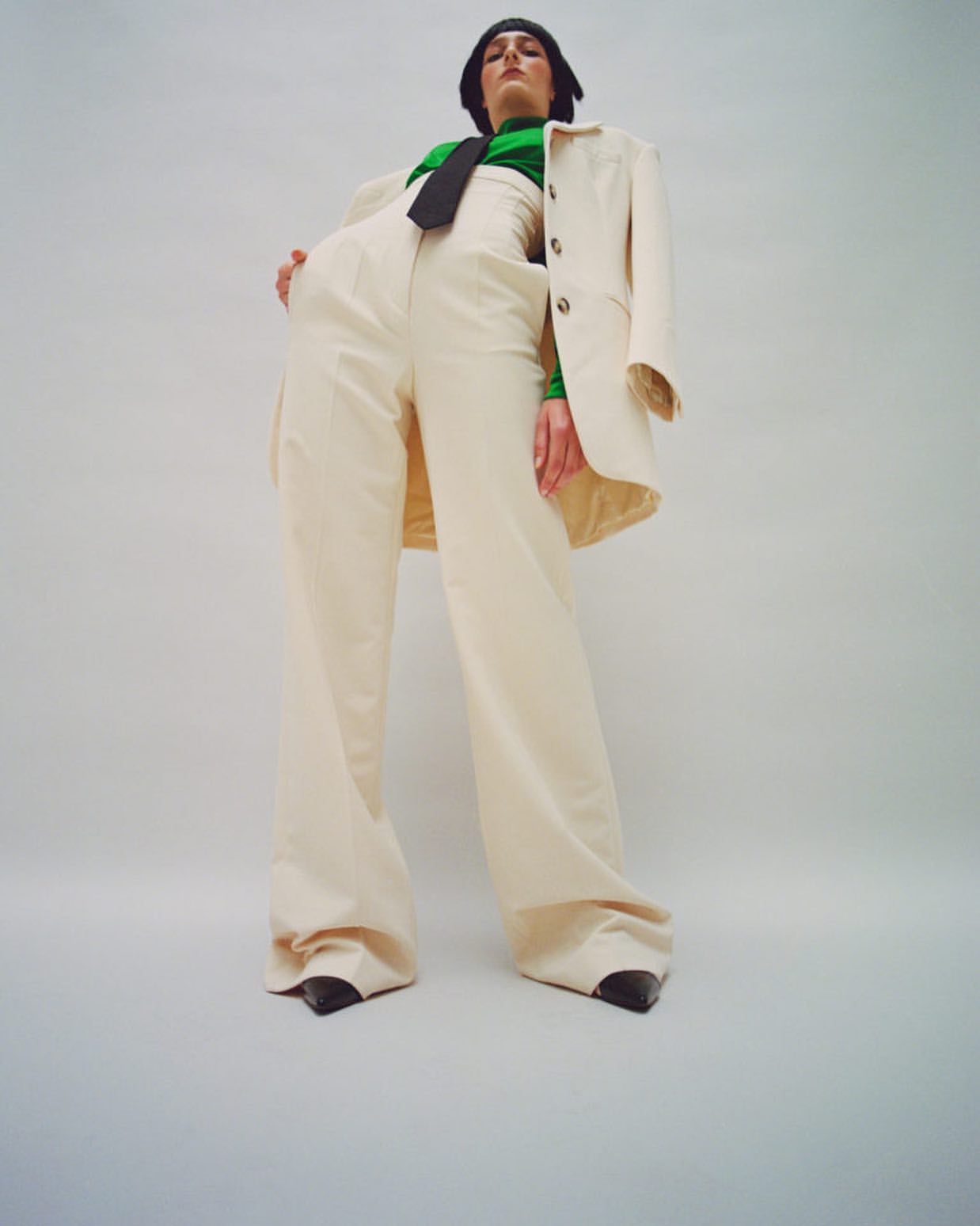 Adult female poses in a beige suit and green turtleneck, shot from a low angle against a bright background in a fashion editorial style.