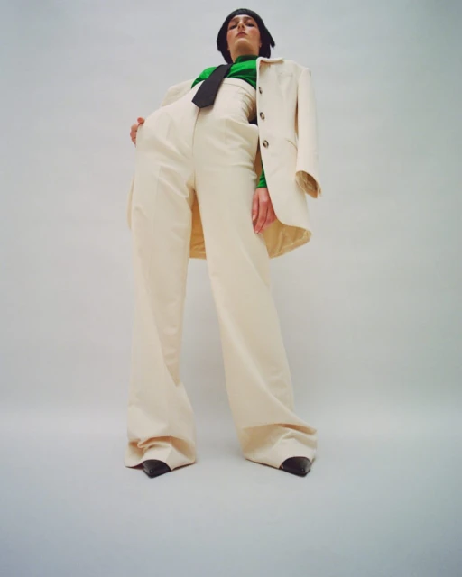 Adult female poses in a beige suit and green turtleneck, shot from a low angle against a bright background in a fashion editorial style.