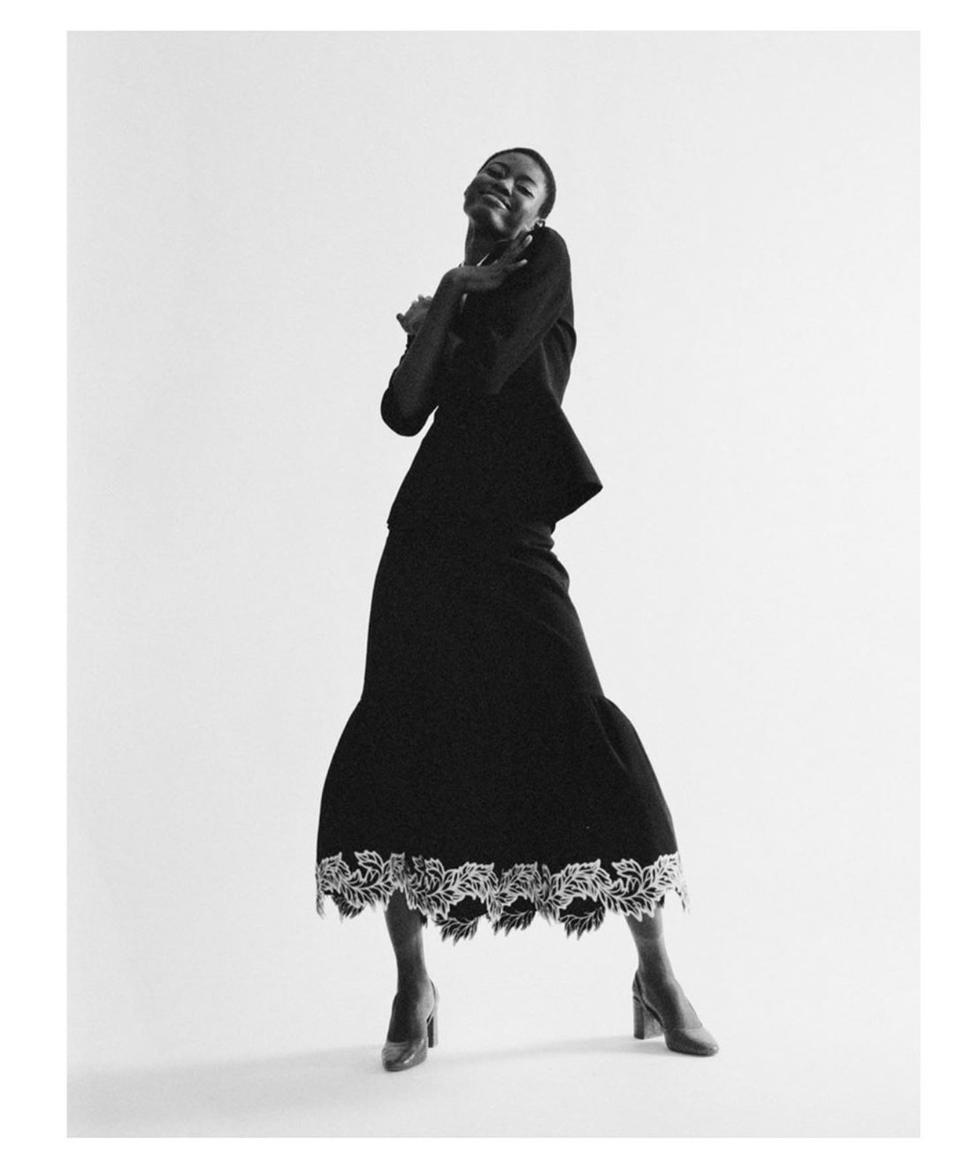A fashionable Black woman stands in a studio wearing a black dress with white floral trim and matching heels against a white background.