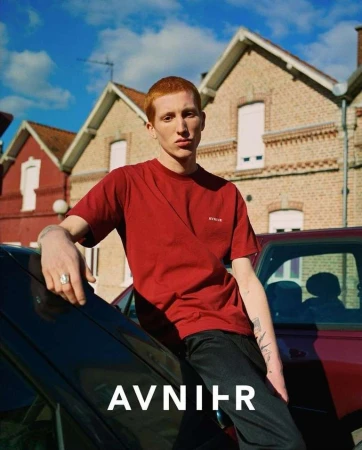 A red-haired man in a burgundy t-shirt leans on a black car in an urban setting with brick houses and a cloudy blue sky.