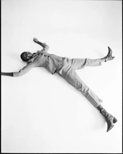 A woman in a grey suit and brown western boots lies in a star pose on a white background in a studio shot.
