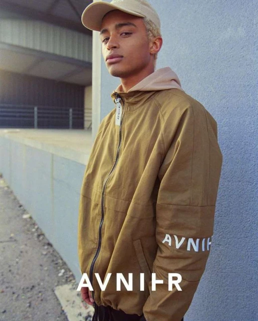 Adult male in a tan jacket and cap poses in front of a concrete wall. Fashion-forward street style with neutral color palette and clean lines.