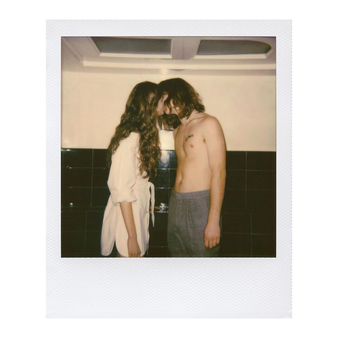 Romantic polaroid-style photograph of a couple embracing in front of dark-tiled walls, featuring one person in a white top.
