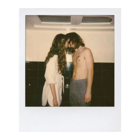 Romantic polaroid-style photograph of a couple embracing in front of dark-tiled walls, featuring one person in a white top.