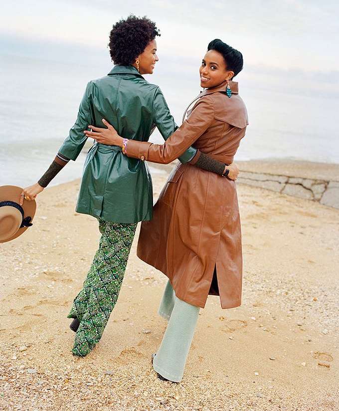 Two women embrace on a sandy beach with ocean view; one wears a green coat and patterned pants, the other a brown coat and blue pants.