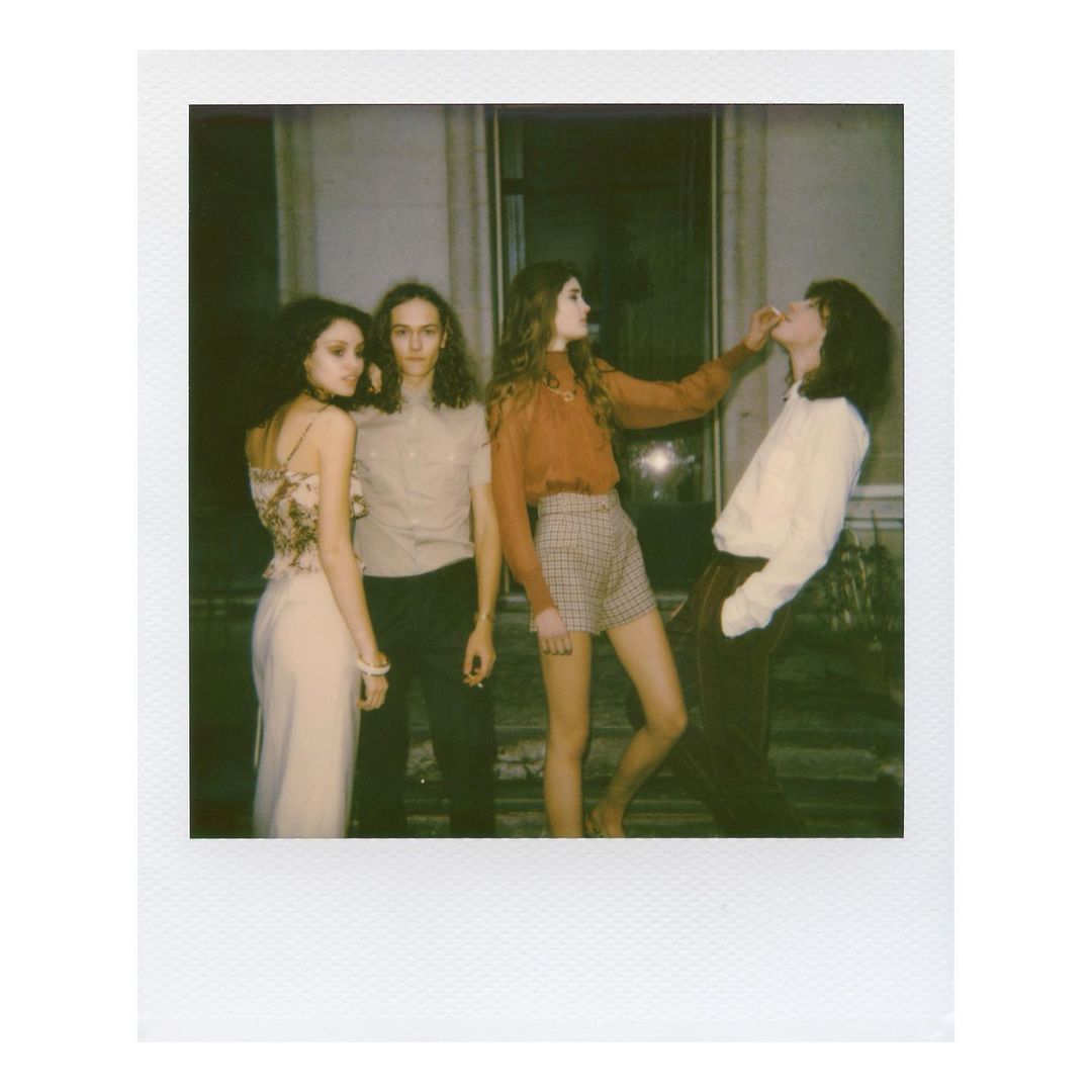 Group of four people in vintage clothing indoors, one woman feeding another, captured in a Polaroid style, with a neutral color palette.