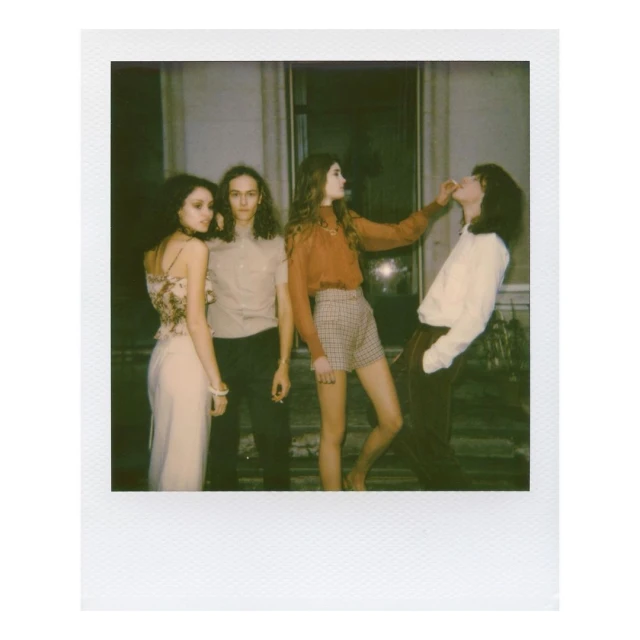Group of four people in vintage clothing indoors, one woman feeding another, captured in a Polaroid style, with a neutral color palette.