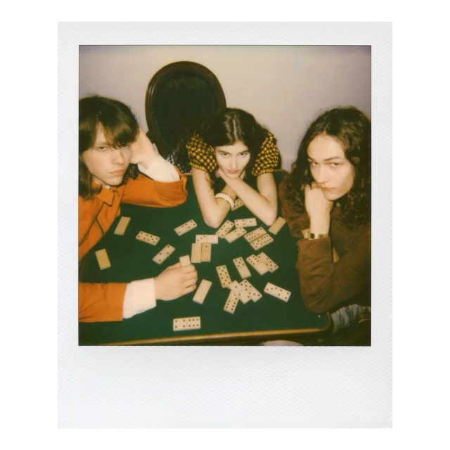 Three people sit at a green table with dominoes. Two women flank a man. They are indoors with a dark oval frame behind them.