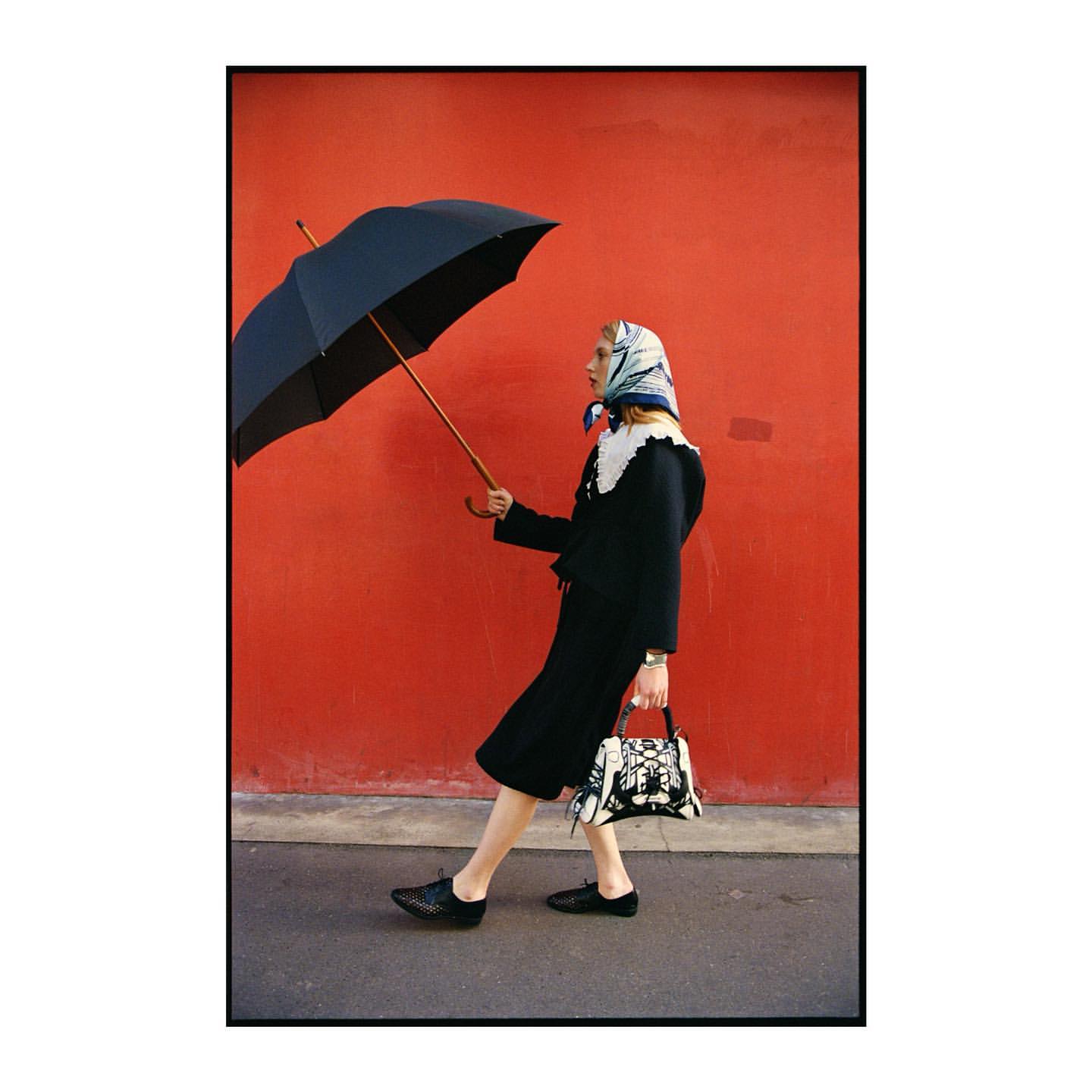 Woman in vintage fashion, wearing a headscarf, dark jacket and skirt, black shoes, and holding an umbrella and patterned handbag.