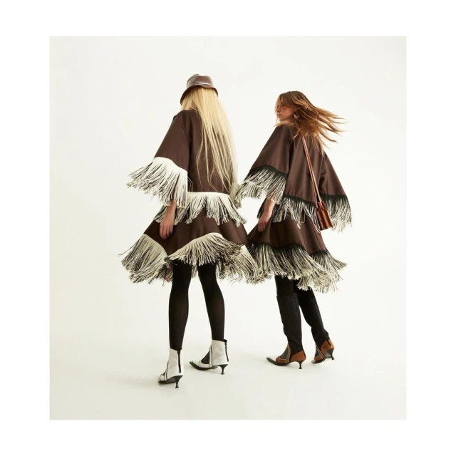 Two women wearing fringed dresses and boots stand against a white background in a fashion forward pose.