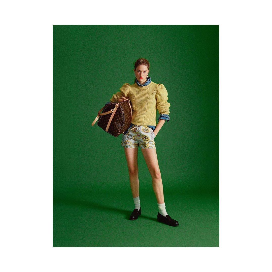 A woman stands in front of a green background, wearing shorts, a sweater, and carrying a brown duffel bag in a fashion photography setting.