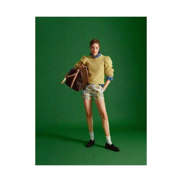 A woman stands in front of a green background, wearing shorts, a sweater, and carrying a brown duffel bag in a fashion photography setting.