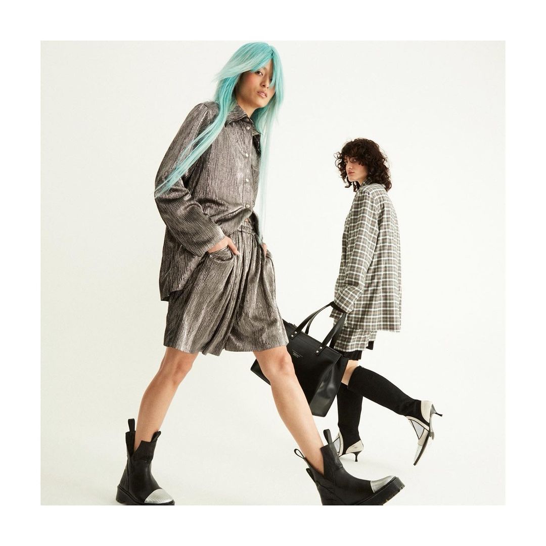 Two adult women pose in a studio wearing metallic-look outfits and black boots against a bright white backdrop in a fashion editorial.