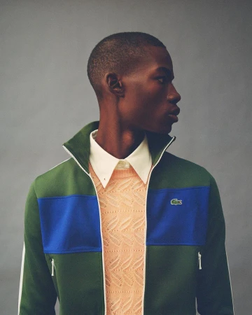 Portrait of a Black man in profile wearing a green track jacket with blue panels over a white collared shirt and peach knit sweater