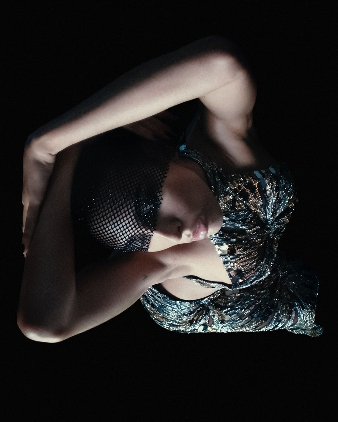 Woman in sparkling top poses in a dark studio. Beauty shot with eye-level angle highlighting her makeup and detailed attire.