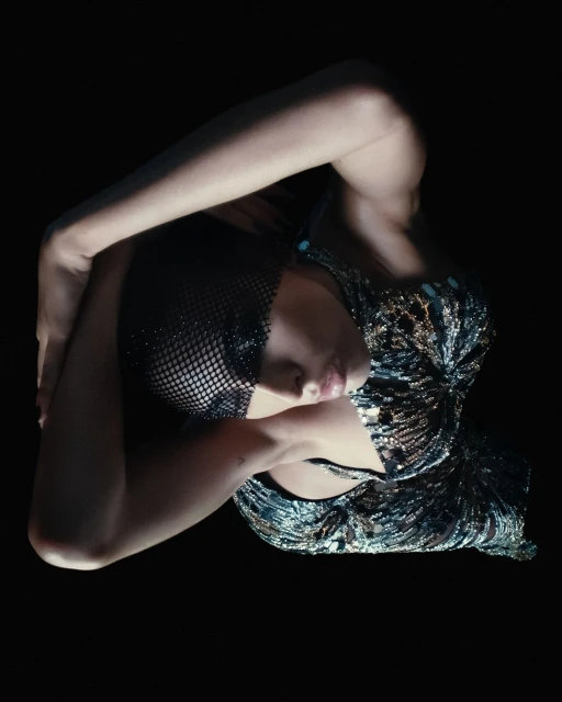 Woman in sparkling top poses in a dark studio. Beauty shot with eye-level angle highlighting her makeup and detailed attire.
