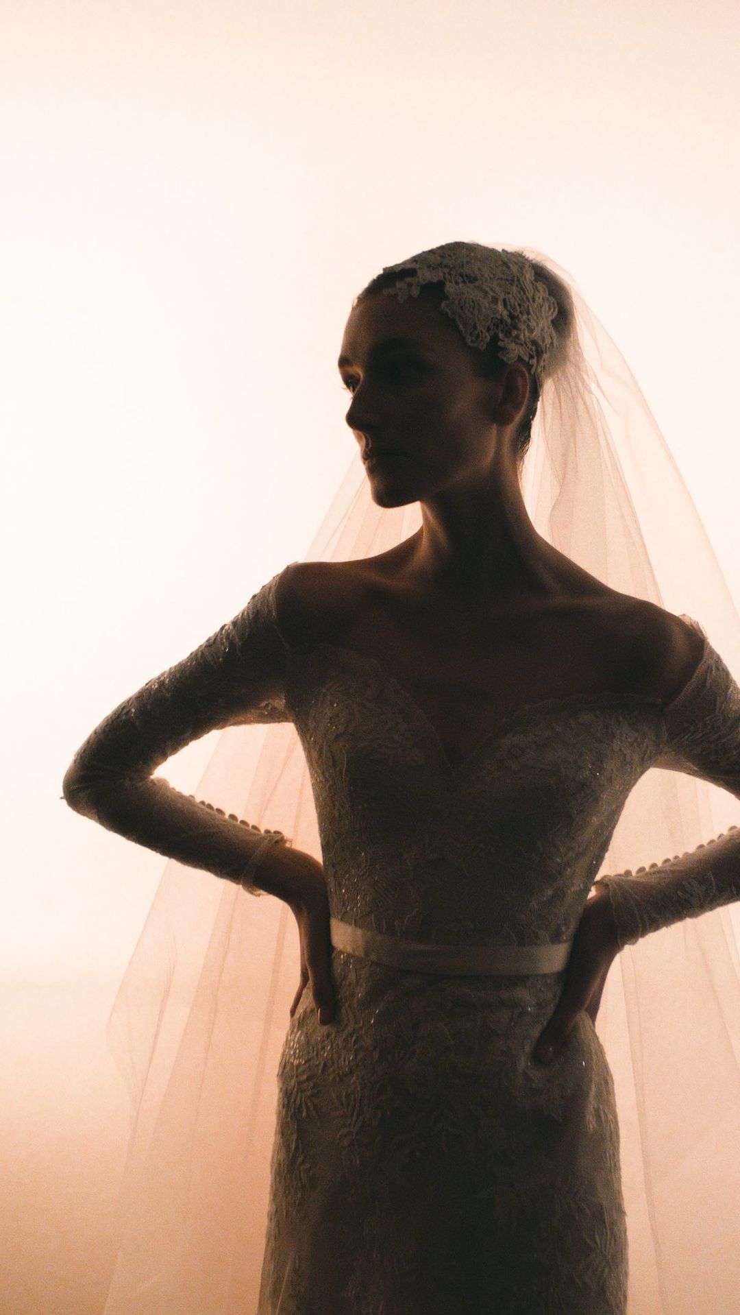 A bride in a long-sleeved beaded gown and veil stands against a gradient backdrop, hands on hips, in a fashion forward silhouette.