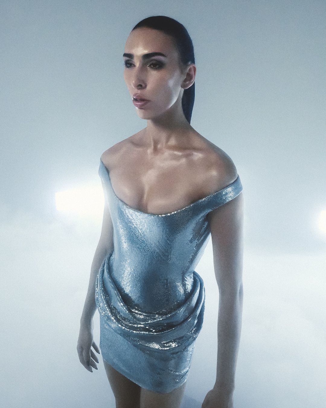 Elegant adult woman in a silver metallic off-the-shoulder dress with cinched waist, posing against a white background with studio lighting.