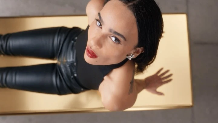 High-angle portrait of Zoe Kravitz wearing a black top and leather pants, seated on a reflective gold surface.